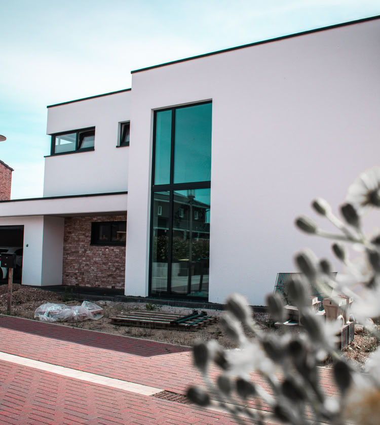 Modernes zweistöckiges Haus mit weißer Fassade, großen Fenstern und einem Carport, in dem ein weißes Auto steht. Im Vordergrund sind unscharfe Pflanzen zu sehen.