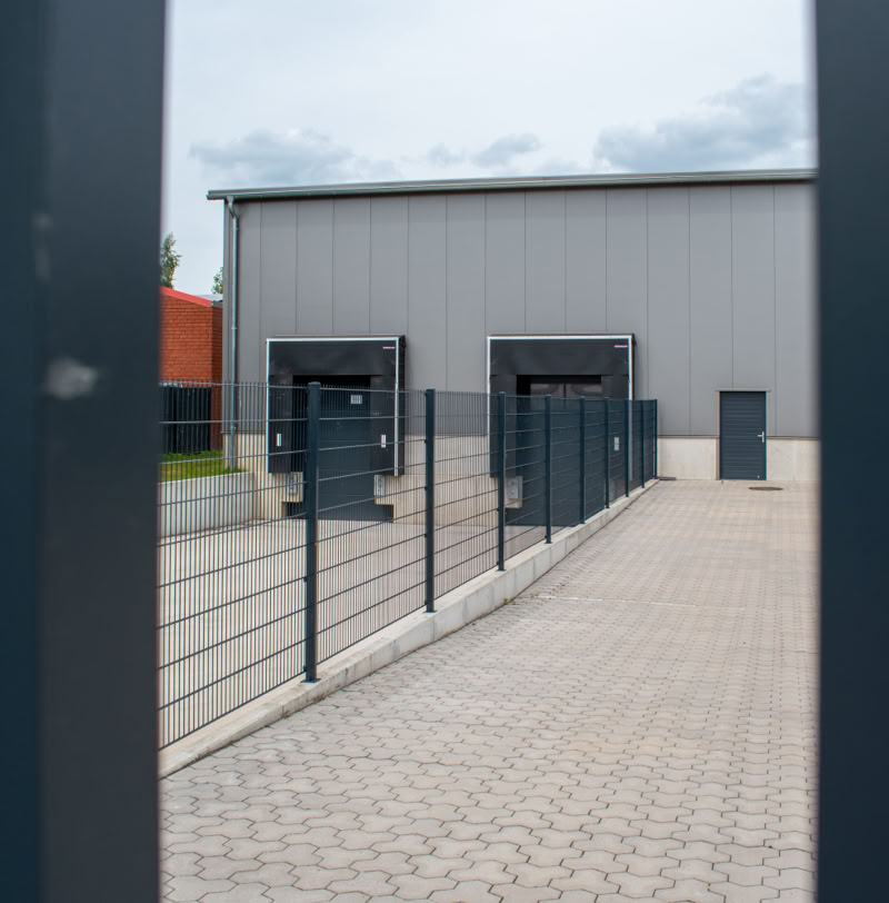 Blick durch einen Zaun auf ein Industriegebäude mit Laderampen und gepflastertem Hof.