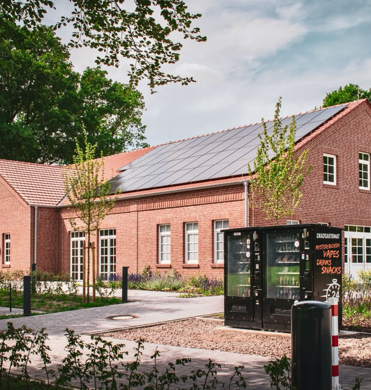 Backsteingebäude mit Satteldach, umgeben von Bäumen und einem gepflasterten Weg im Vordergrund.