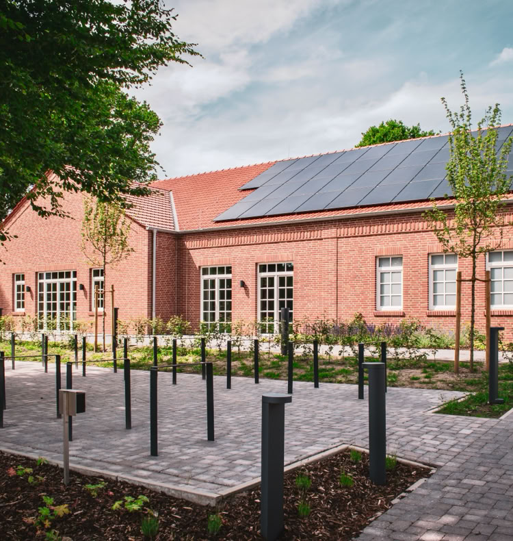 Backsteingebäude mit Solarpanelen auf dem Dach, umgeben von einem gepflasterten Weg und Bäumen.