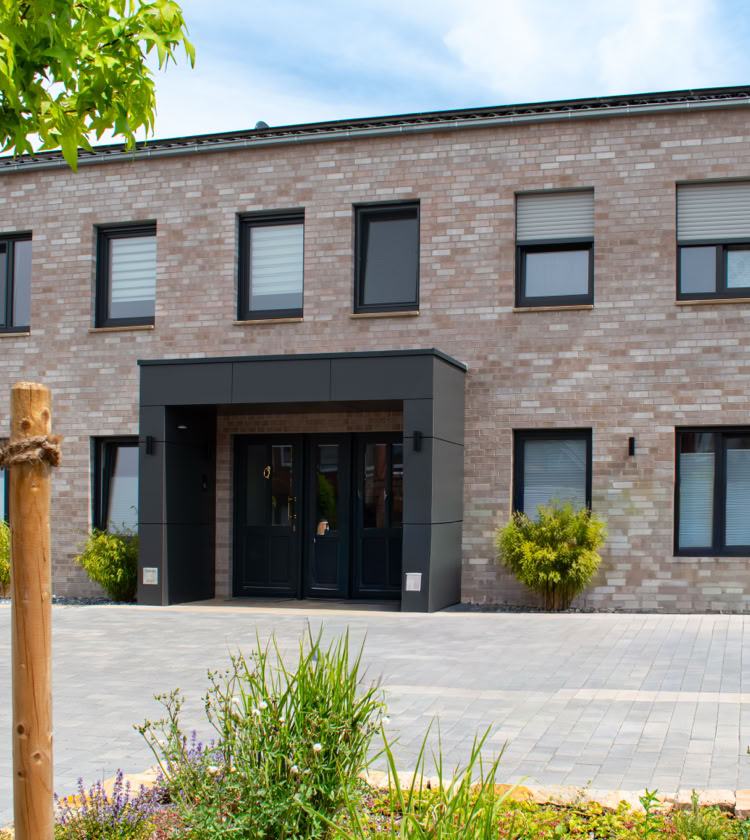 Zweistöckiges Gebäude aus Ziegelsteinen mit mehreren Fenstern und einer Eingangstür, umgeben von Pflanzen und einem gepflasterten Weg.