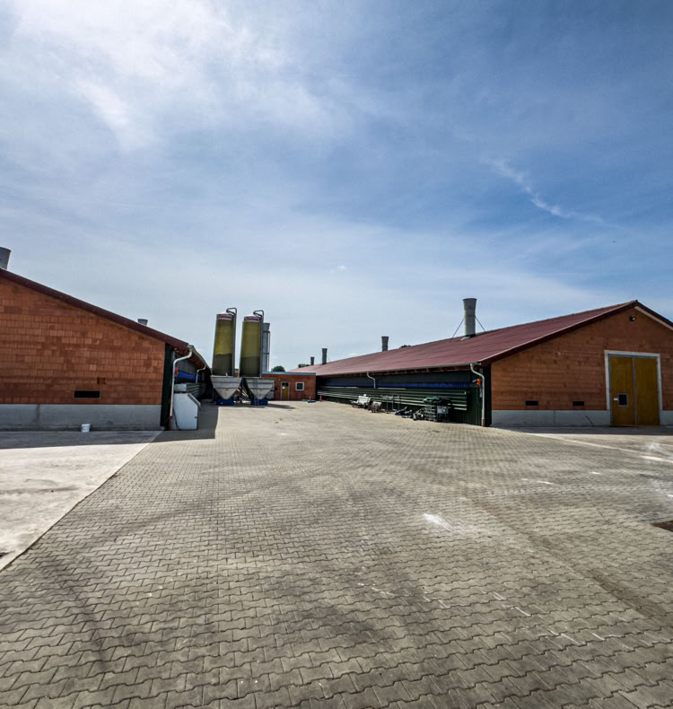 Zwei große, rote Backsteingebäude mit gelben Türen auf einem gepflasterten Hof, im Hintergrund zwei Silos und blauer Himmel.