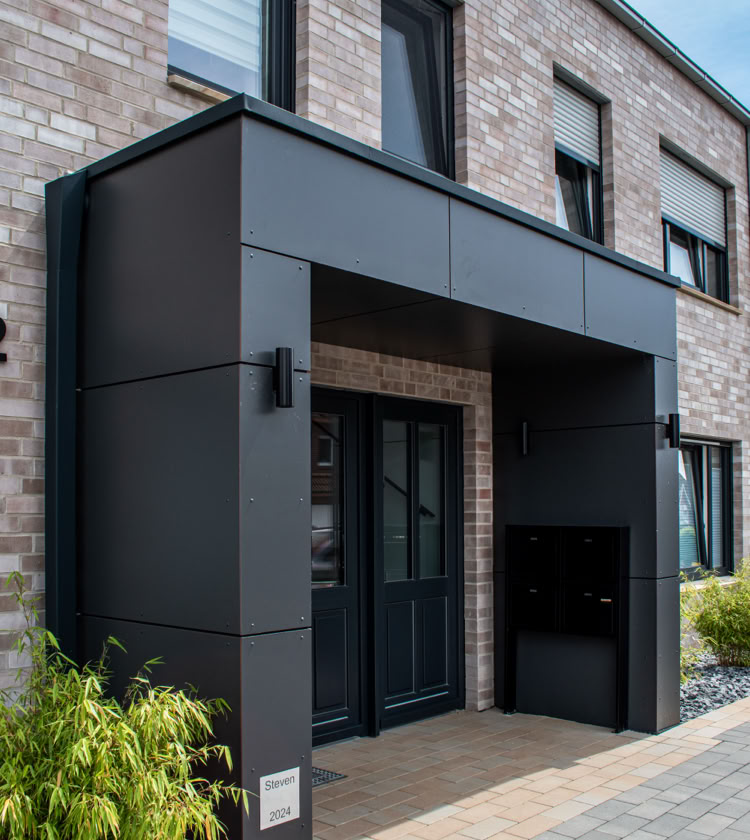 Moderne Hausfassade aus Backstein mit einem schwarzen Vordach über dem Eingang, flankiert von Fenstern und Pflanzen.