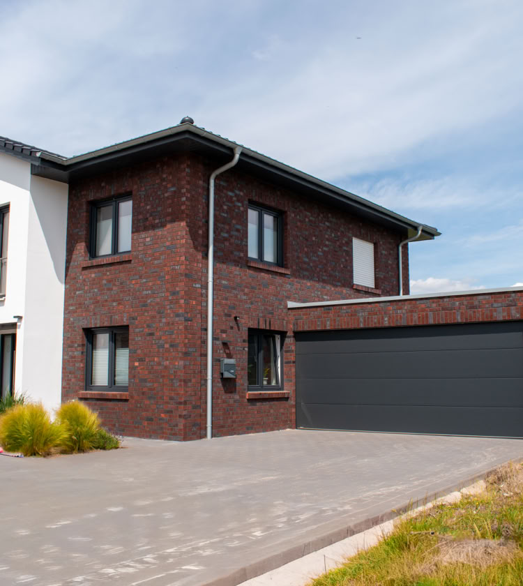 Zweistöckiges modernes Haus mit roter Backsteinfassade, weißen Akzenten und einer großen Garage, umgeben von einem gepflasterten Hof und etwas Grünfläche.