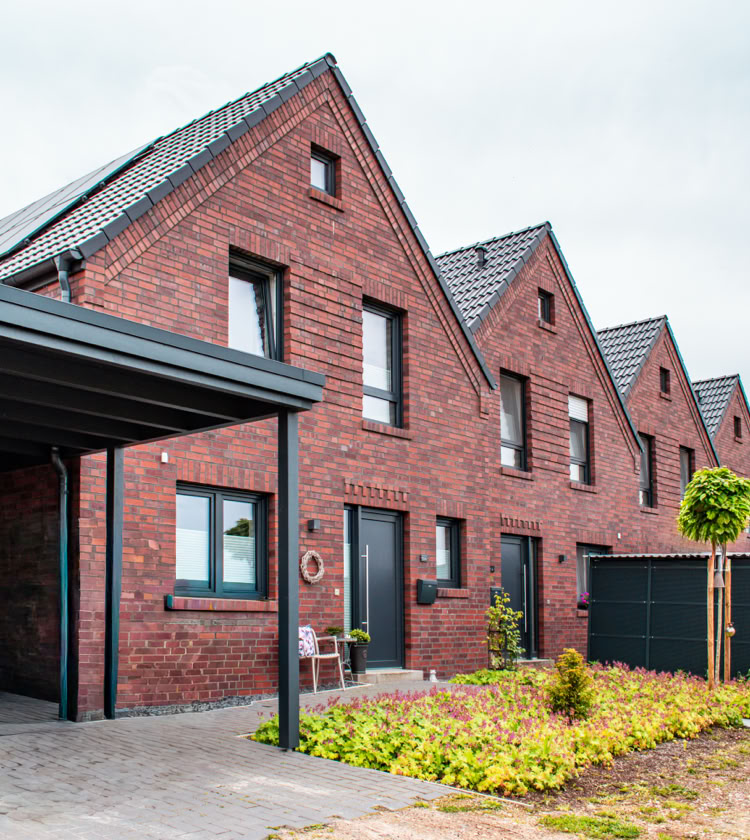 Reihenhäuser aus rotem Backstein mit spitzen Dächern, Vorgärten und einem Carport auf der linken Seite.